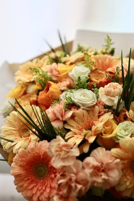 A vibrant floral bouquet featuring a mix of soft peach and cream-colored gerbera daisies, roses, and spray roses, accented with delicate green foliage and long, slender palm leaves. The flowers are ar