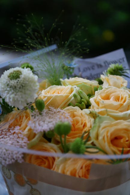 A close-up of a floral arrangement featuring soft peach roses with gently unfurling petals, complemented by small white and green blooms, including fluffy white pom-pom-like flowers and tiny green bud