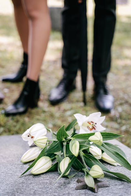 A floral arrangement of white lilies with large, open petals and prominent stamens, accompanied by several closed white lily buds and green leaves with elongated shapes. The flowers are laid on a ston