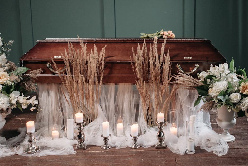 A floral arrangement for a funeral comprising white roses, lilies, and other soft-colored blooms in white vases on either side of a polished wooden casket, adorned with beige pampas grass and sheer wh